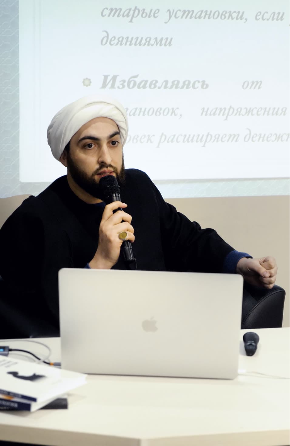 Учитель Хусейн говорит с микрофоном на семинаре, перед ним раскрытый ноутбук
