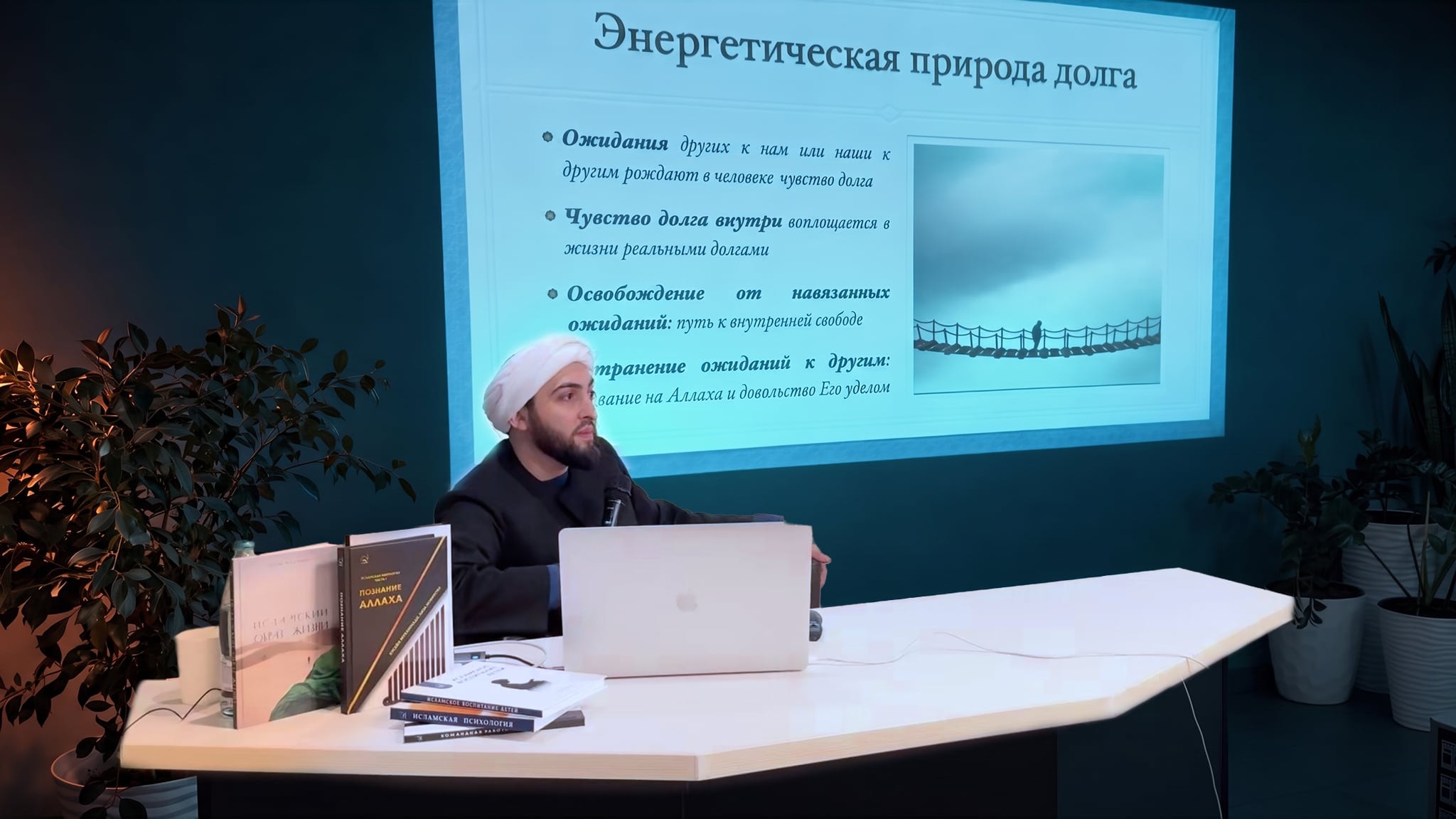 Учитель Хусейн ведёт семинар за столом, на экране за ним слайд об энергетической природе долга
